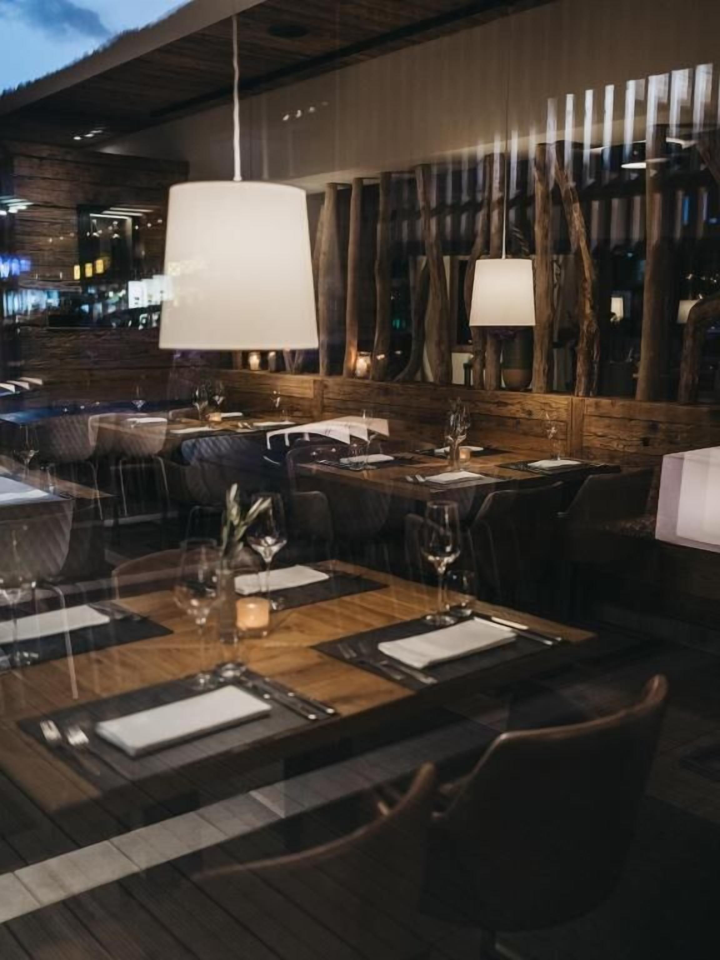 Un restaurante con mesas y sillas de madera, grandes lámparas colgantes y vistas a la ciudad iluminada de noche.