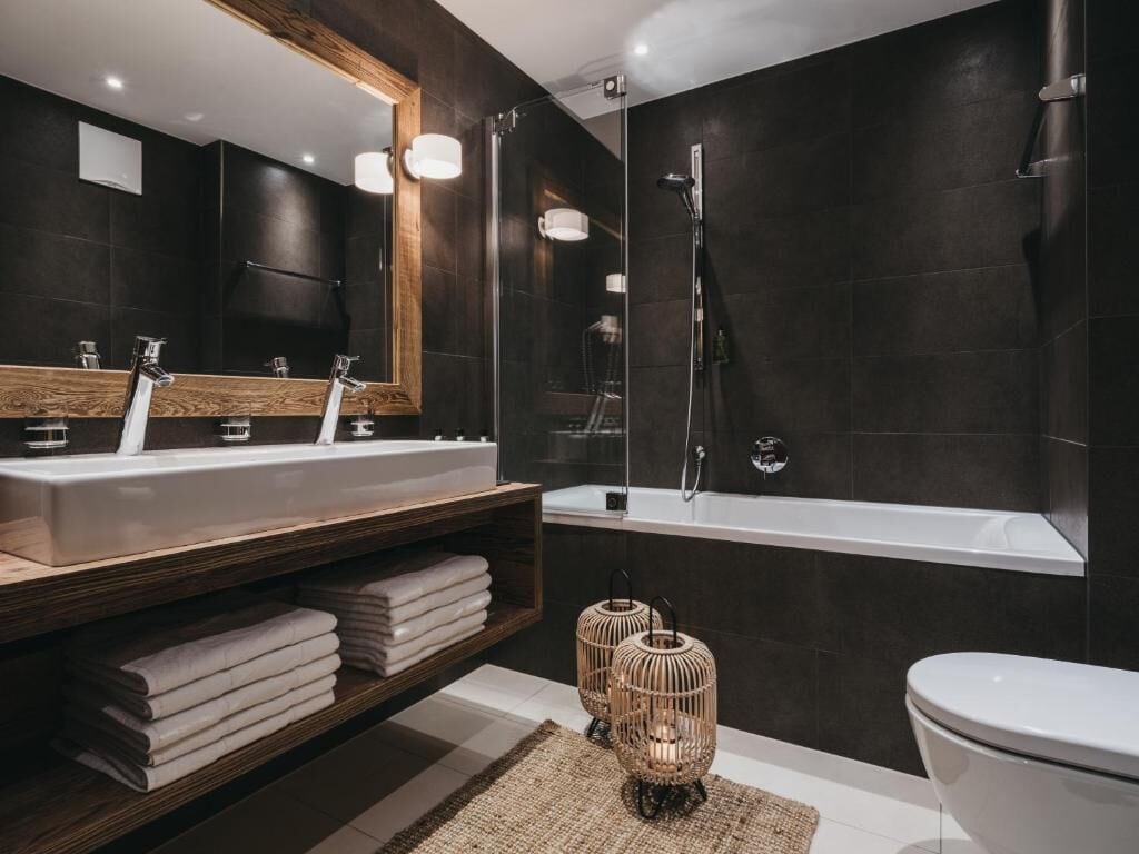 Une salle de bain moderne avec des carreaux foncés, un grand miroir, deux lavabos, une baignoire et des toilettes.