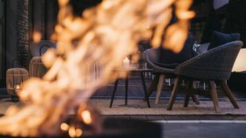 Un rincón al aire libre acogedor con sillas de mimbre y una mesa, una pequeña chimenea al frente y un muro de ladrillo al fondo.