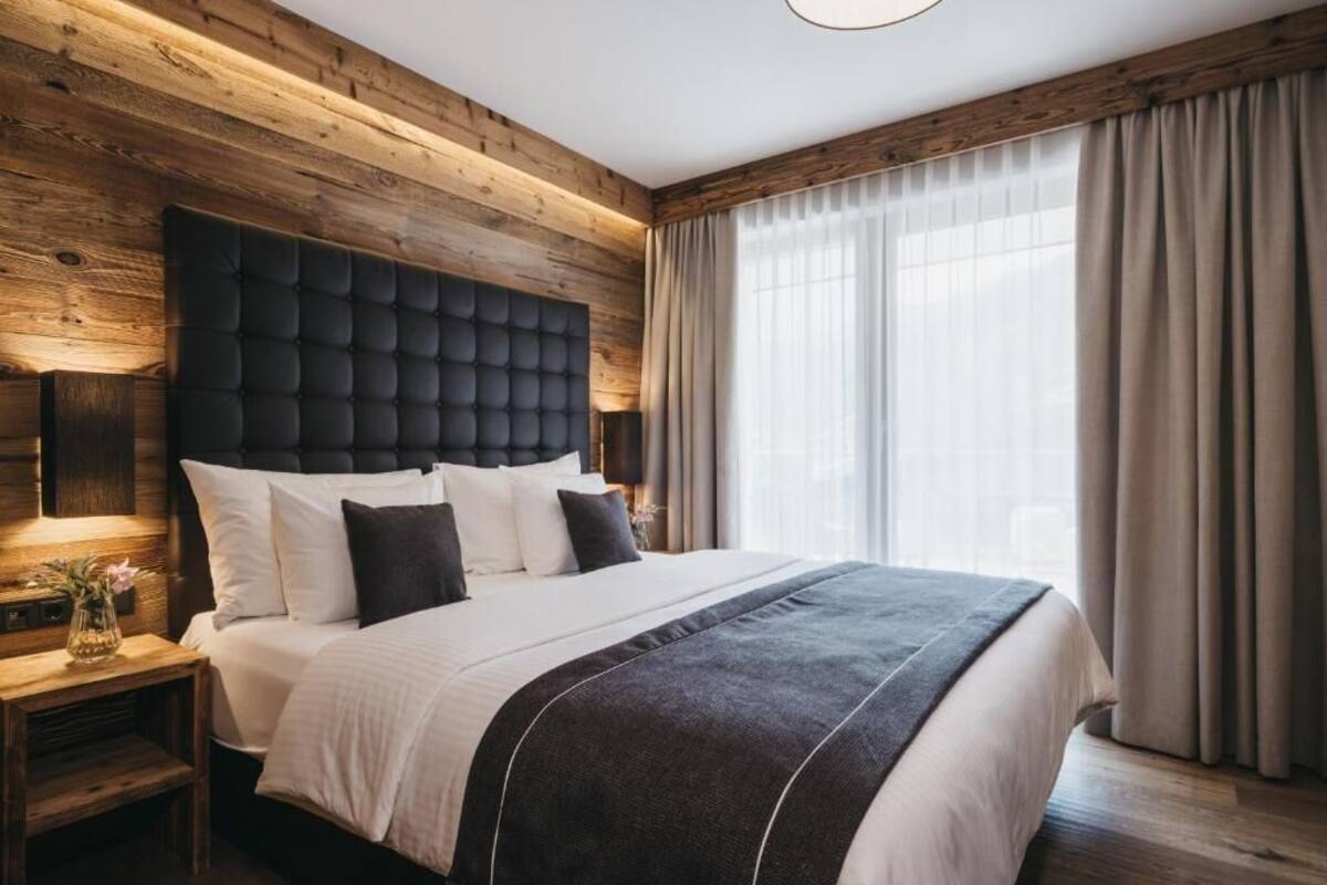 Un hotel moderno con una cabecera de madera, una cama grande con ropa blanca, una mesita de noche con lámpara y vistas al exterior a través de cortinas translúcidas.