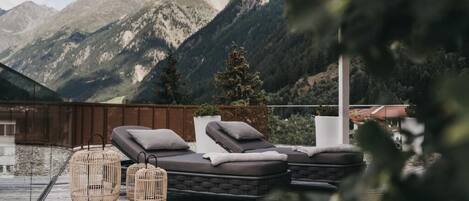 A rooftop terrace with lounge chairs and wicker tables, overlooking a mountainous landscape.