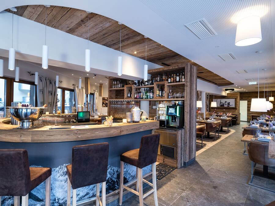A modern bar area with a wooden ceiling, bar stools, and a variety of bottles and glasses on shelves.
