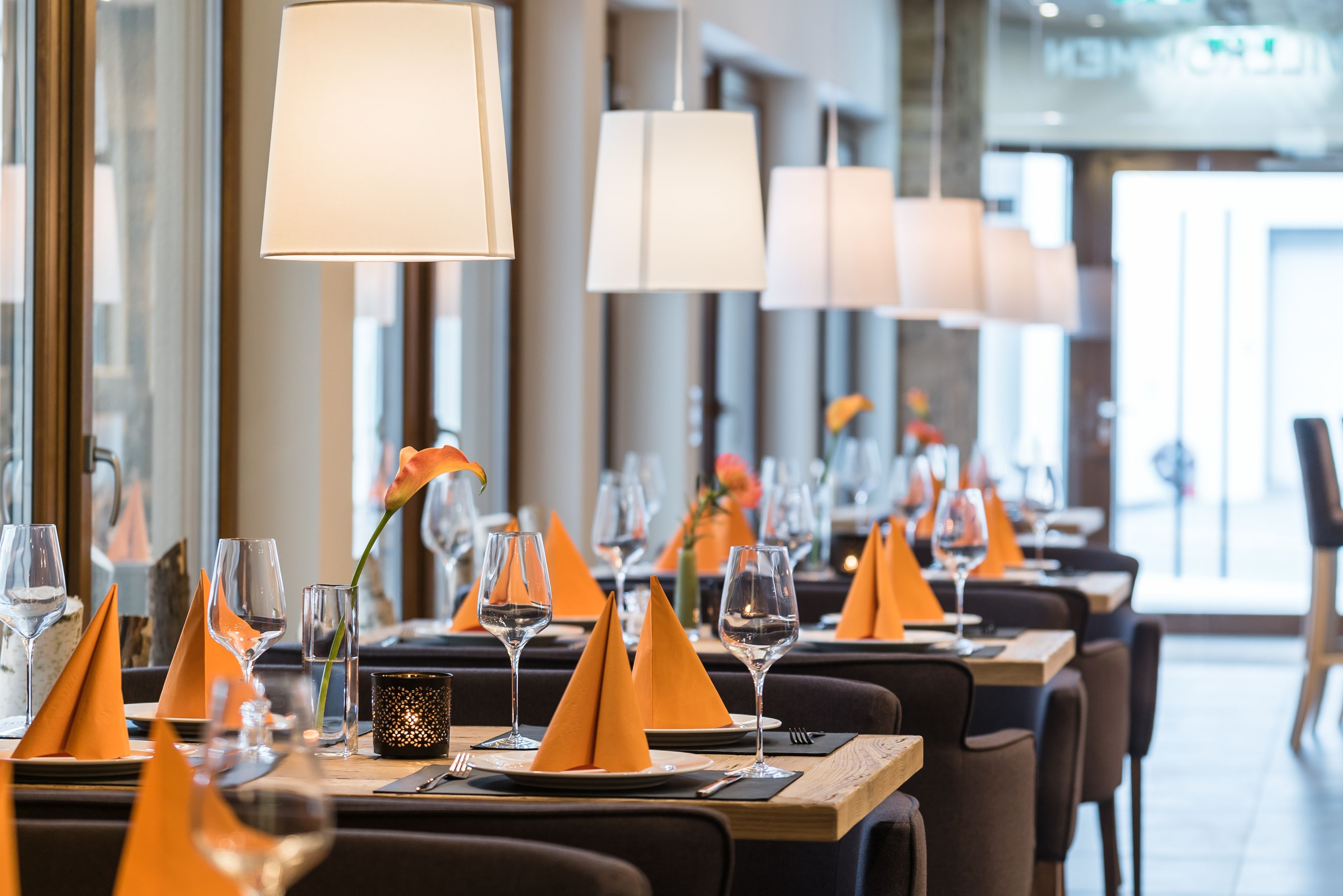 Un restaurant dont les tables sont dressées avec des serviettes orange, des verres à vin et un vase contenant une fleur.