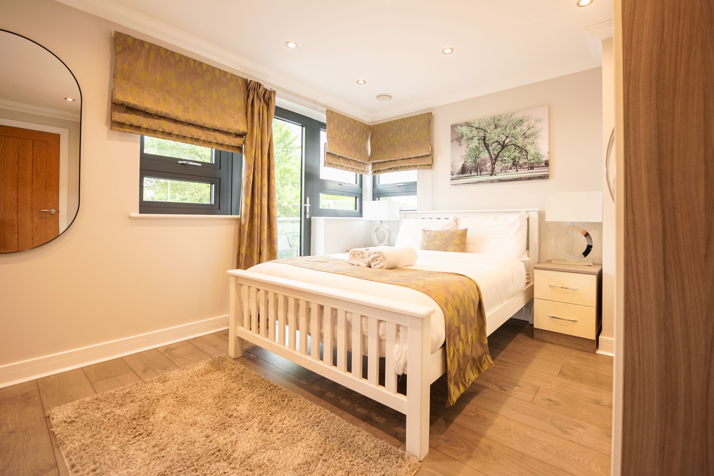Une chambre avec un lit blanc, une table de chevet, une commode, une fenêtre avec des rideaux et un miroir fixé au mur.