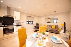 A modern kitchen with a stove, oven, and cabinets, a dining area with yellow chairs and a table set for two, and a living area with a sofa and a coffee table. - Malthouse Court (Reading)