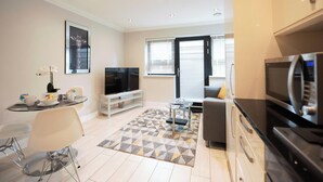 A modern living room with a flat-screen TV, a sofa, a dining area with a glass table, and a kitchen with a refrigerator. - Malthouse Court (Reading)