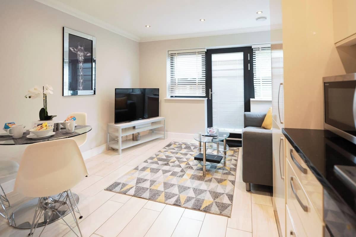 Uma sala de estar moderna com televisão de tela plana, sofá, área de jantar com mesa de vidro e cozinha com geladeira.