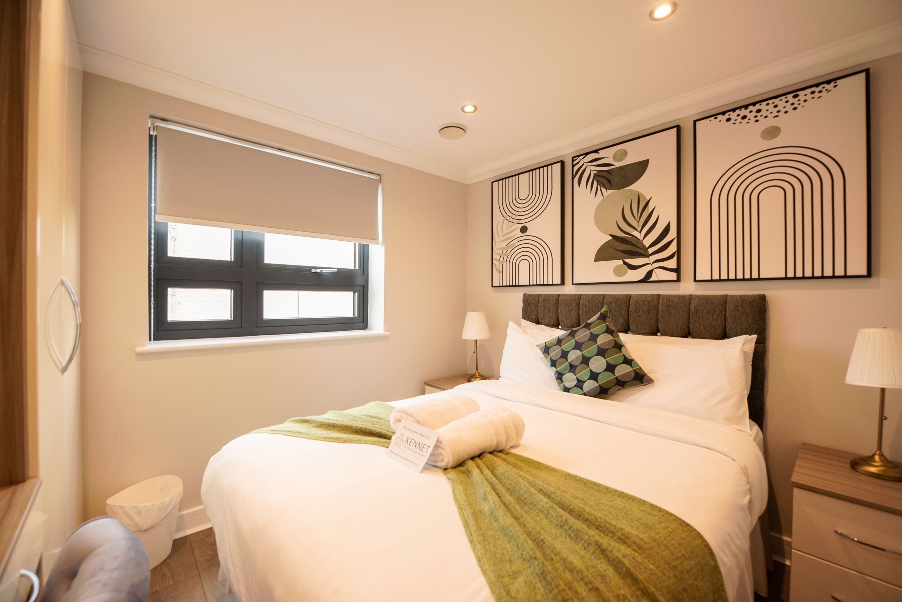 A hotel room with a bed, a window with blinds, a bedside table with a lamp, and wall art.