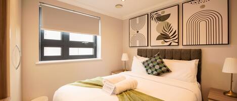Habitación de hotel con cama, ventana con persianas, mesita de noche con lámpara y decoración en la pared.