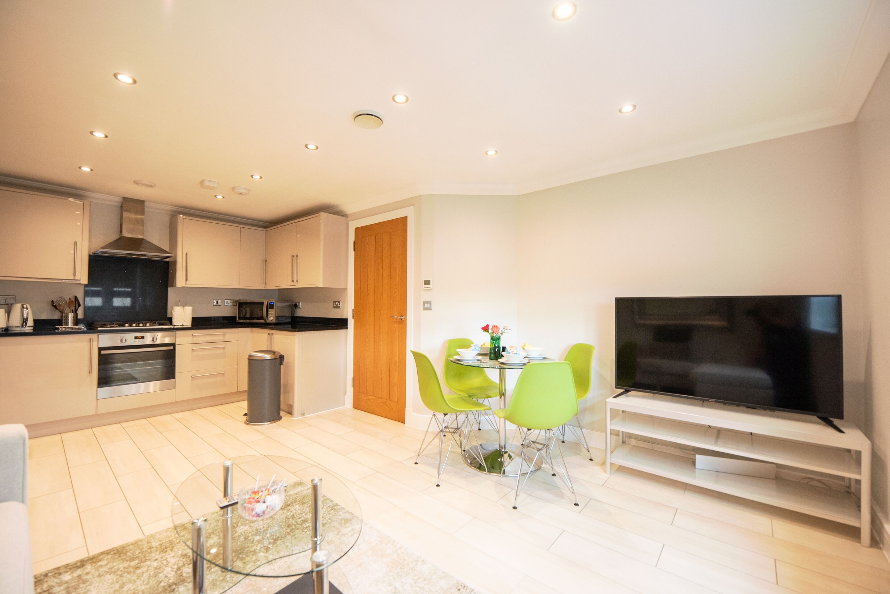 A modern kitchen with built-in appliances, a dining area with green chairs, and a flat-screen TV on a stand.