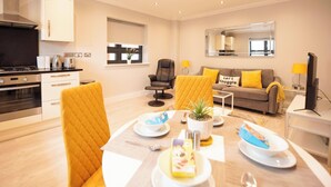 A modern kitchen with a stove, oven, and cabinets. There is a dining area with a table, chairs, and a plant. A sofa and a TV are also visible. - Malthouse Court (Reading)