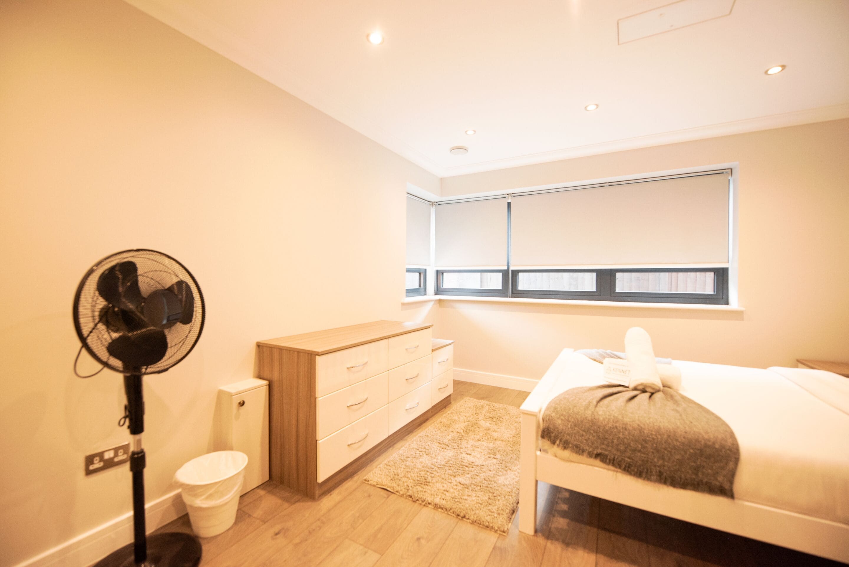 A modern bedroom with a bed, a wooden dresser, a fan, a window, and a small white trash bin.