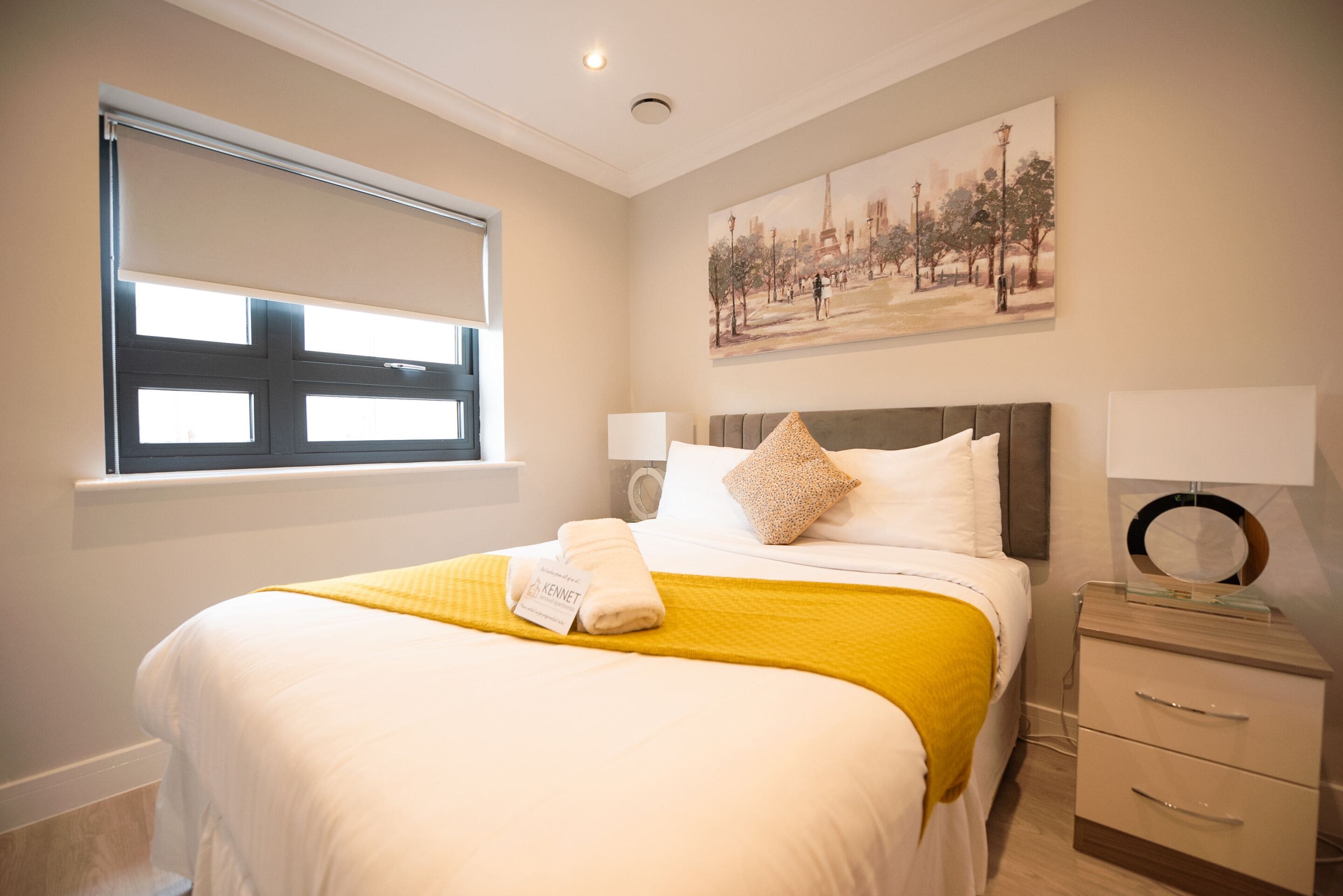 A hotel room with a bed, a window with blinds, a painting on the wall, a bedside table with a lamp, and a small drawer unit.