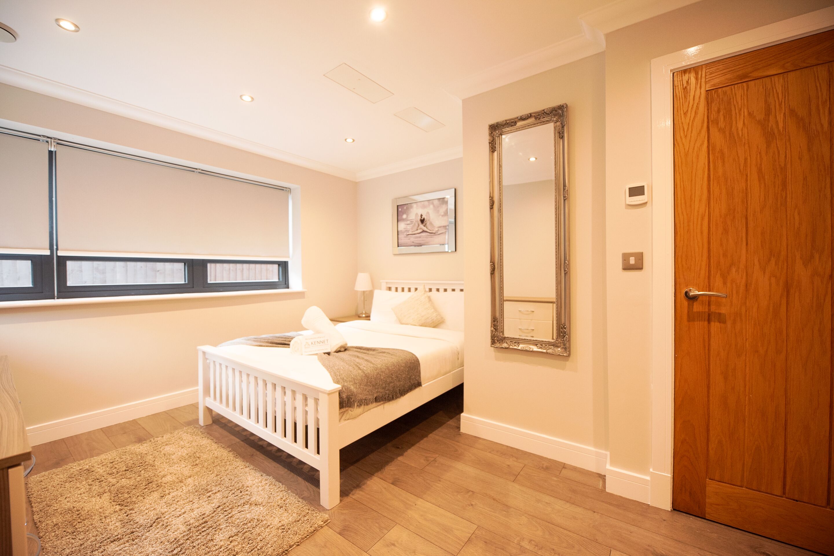 A bedroom with a white bed, a large mirror, a wooden door, and a window with blinds.