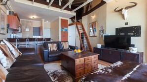 A spacious living room with a leather sofa, a wooden coffee table, and a television. A large bull skull decorates the wall, and a staircase leads to an upper level. - Leisure Bay 306 by CTHA (Cape Town)