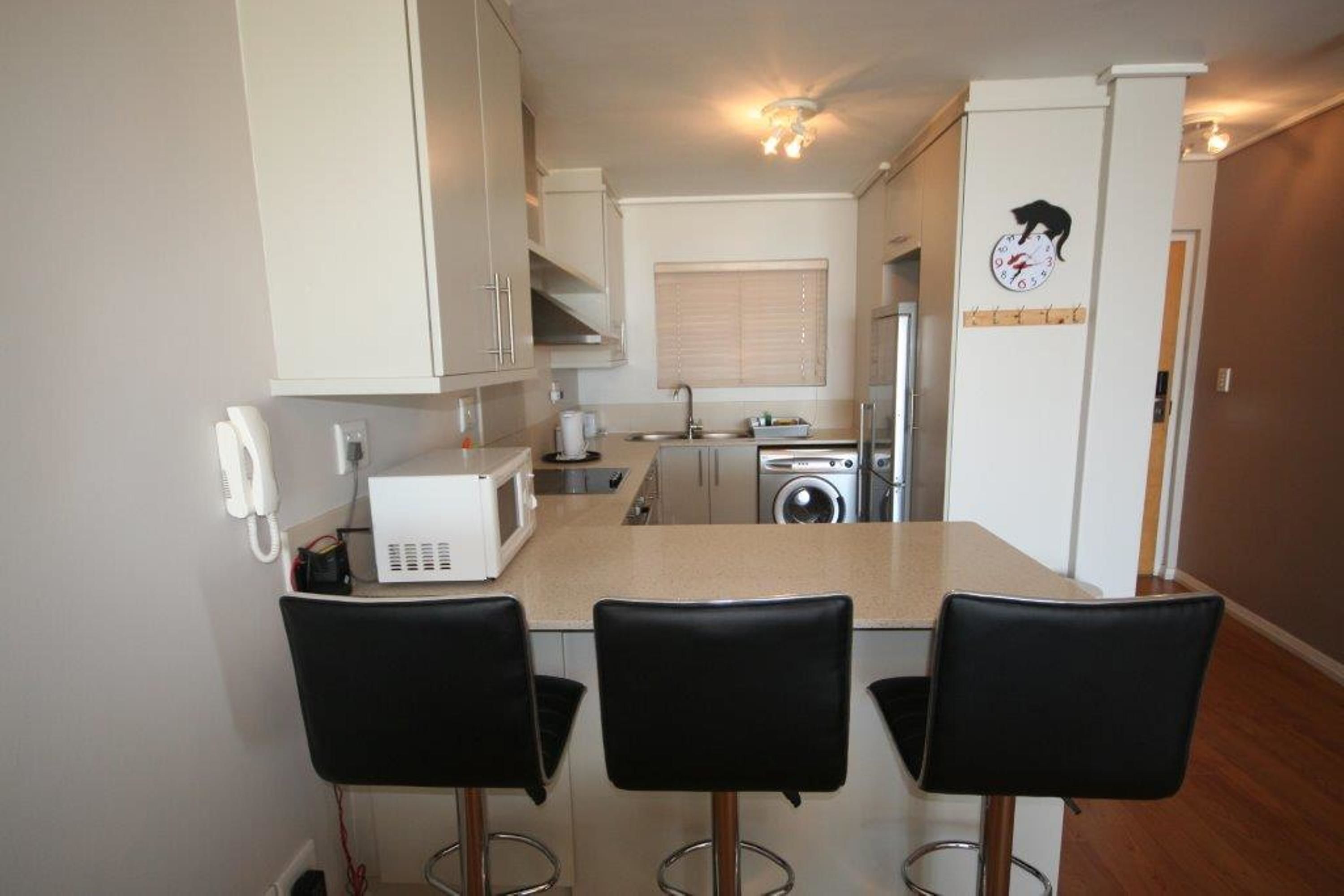 A compact kitchen with a microwave, toaster, sink, and washing machine. There are three black bar stools and a white telephone.