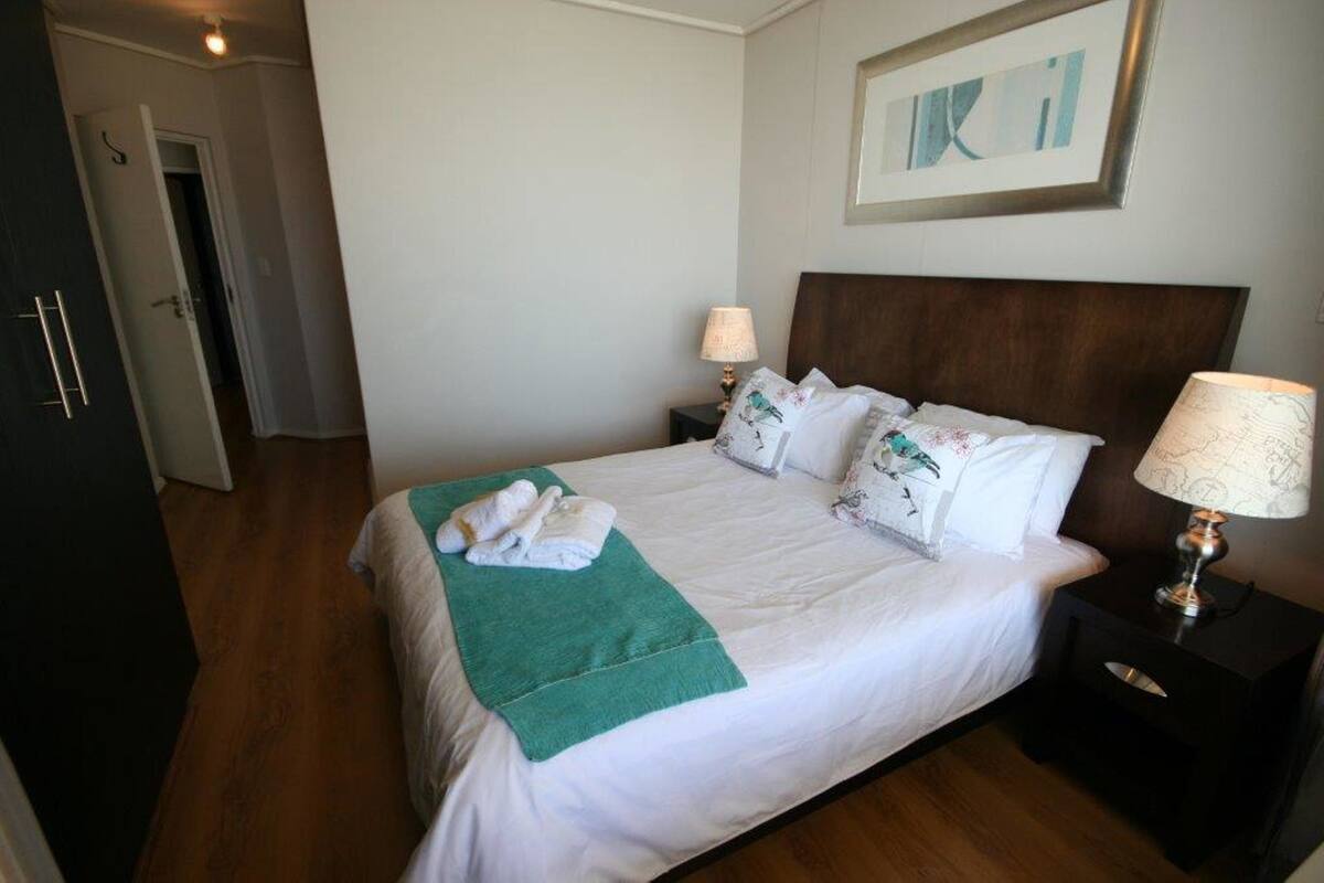Een netjes opgemaakt bed met witte lakens en een groene plaid, twee nachtlampjes en een ingelijst kunstwerk aan de muur.
