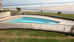 Une piscine dotée d’une terrasse incurvée, entourée d’une clôture métallique, et offrant une vue sur une plage où se trouvent un banc et un lampadaire.