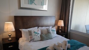 Un lit bien fait, agrémenté d’un plaid turquoise et de plusieurs coussins, dont certains arborent des motifs floraux. Deux lampes de chevet sont placées de chaque côté du lit.