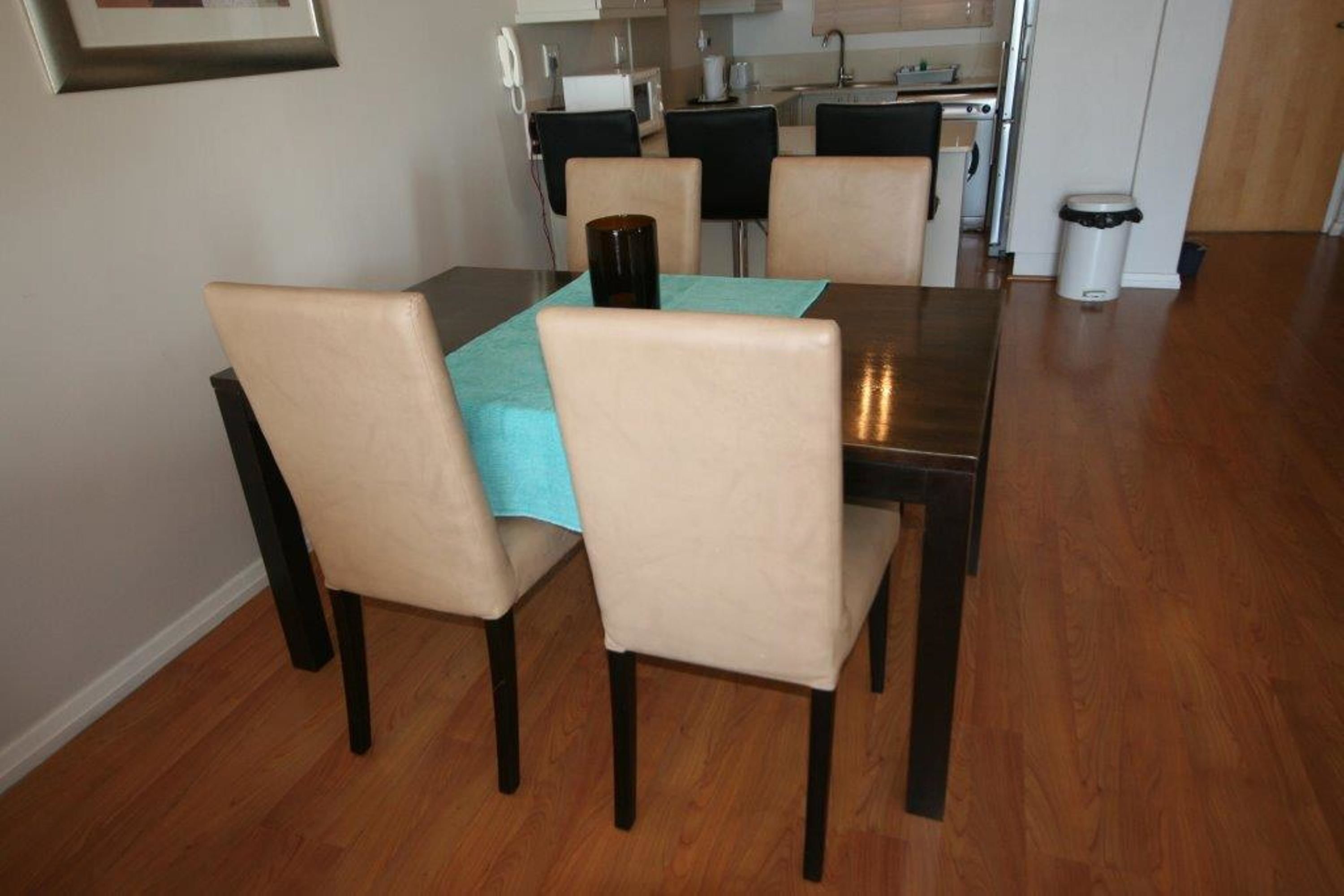 A dining area with a wooden table and four chairs. A trash can is visible in the background.