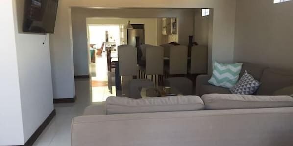 A living room with a beige sofa, a television mounted on the wall, and a dining area in the background.