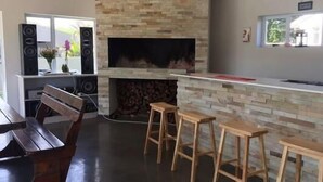 A modern kitchen with a stone backsplash, a built-in fireplace, wooden bar stools, and a dining area with a bench and table. - Clairwood Villa (Cape Town)