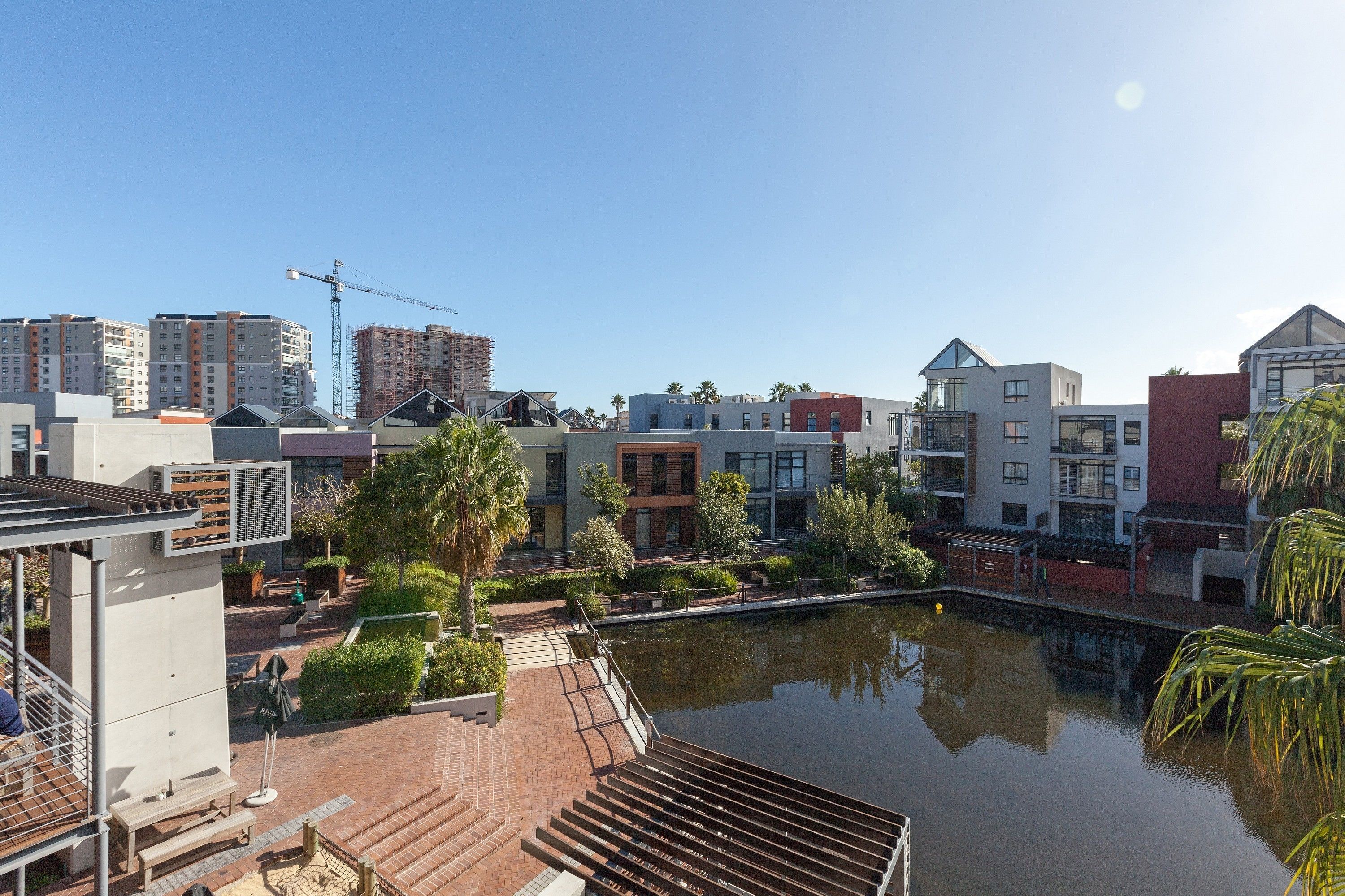 A modern urban area with a calm water body, contemporary buildings, and a construction crane in the background.