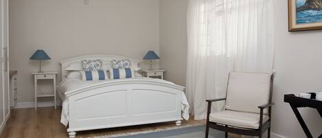 A bedroom with a white bed, two bedside tables with lamps, a chair, and a large window with curtains.