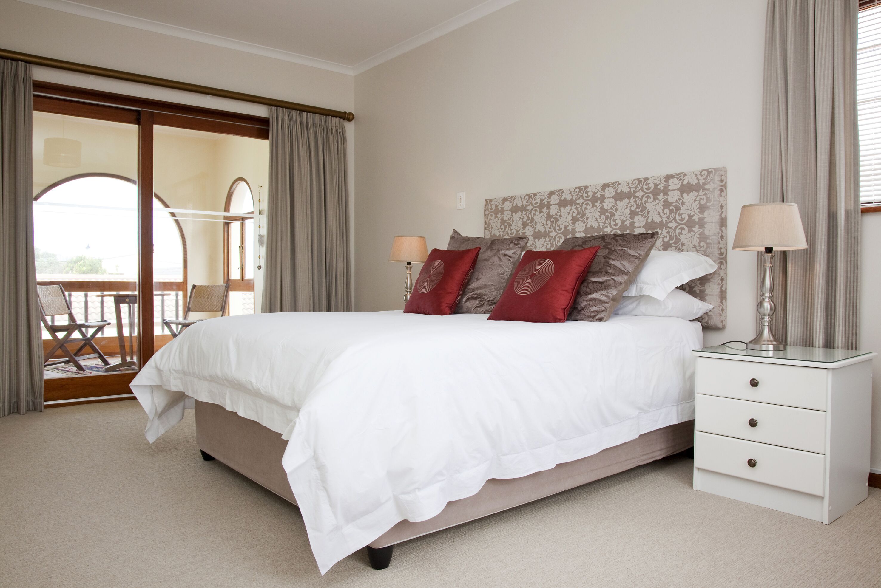 Una cama bien hecha con ropa de cama blanca y almohadas rojas y marrones, una mesita de noche con lámparas, y vistas a un balcón con sillas.