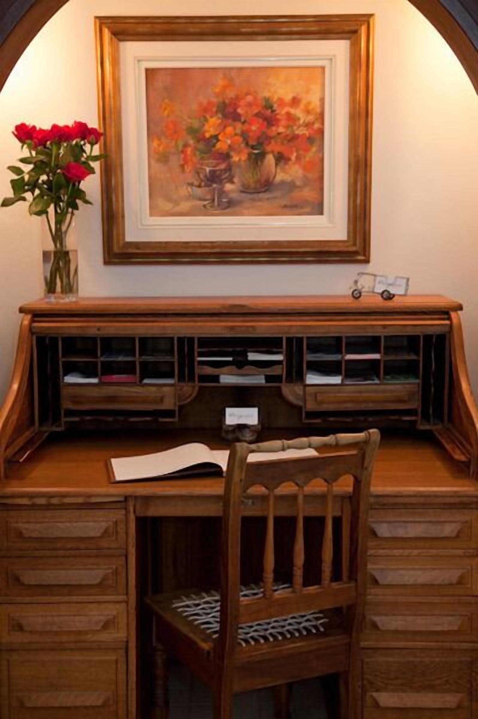 A wooden desk with a chair, a vase with red flowers, and a framed painting above the desk.