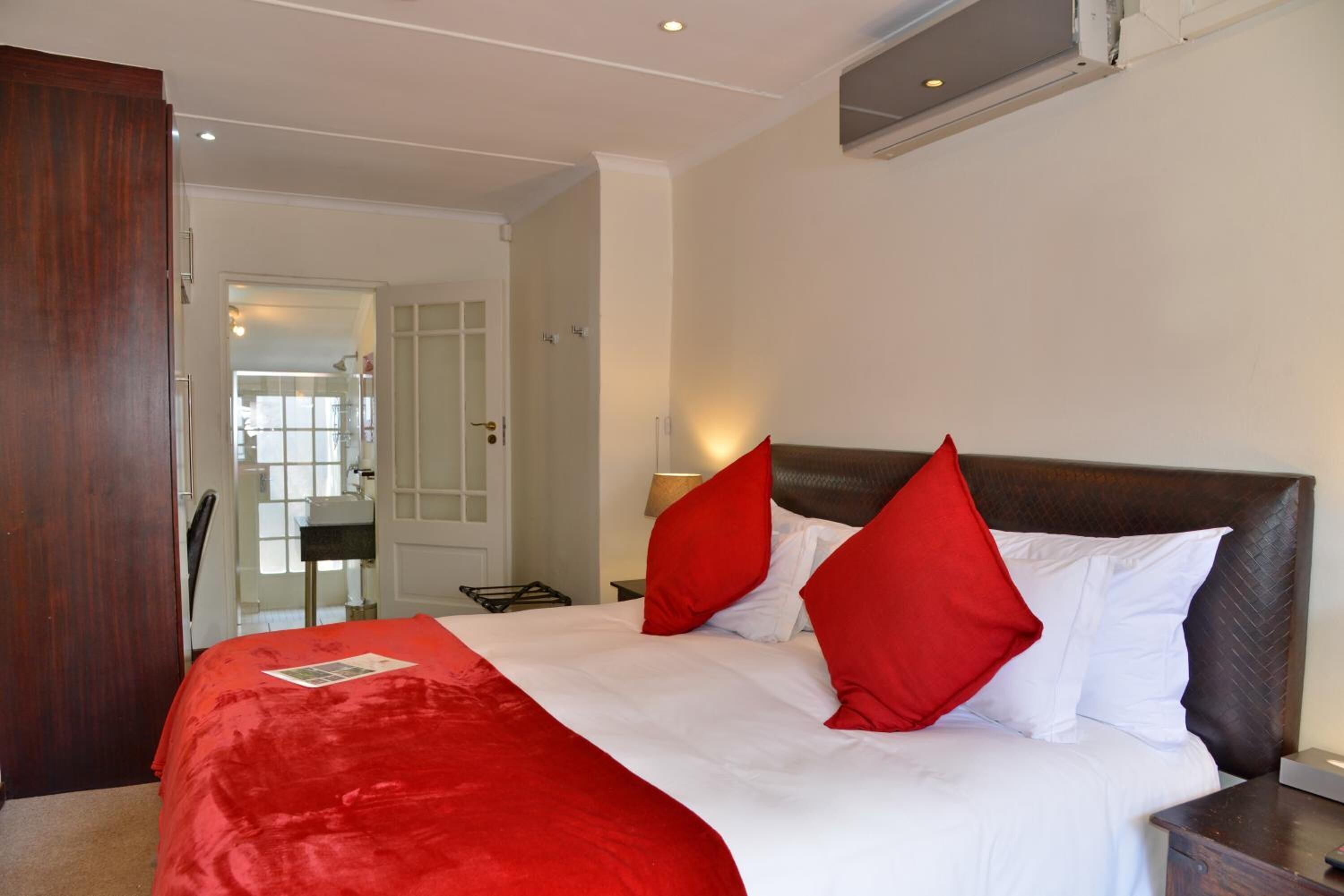 Habitación de hotel con una cama grande, ropa de cama roja y blanca, un cabecero de madera y una mesita de noche con una lámpara.