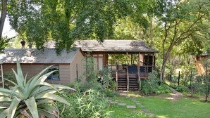 A rustic cabin with a wooden porch, surrounded by lush greenery and a stone pathway. - Sand River Guest House (Sandton)