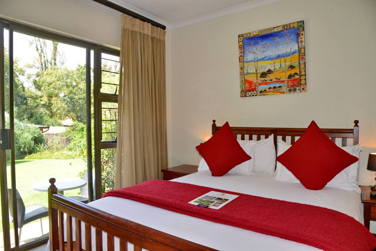 A hotel room with a large bed, red pillows, a red blanket, a wooden headboard, a nightstand, a table, and a painting on the wall.