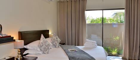 Habitación de hotel con una cama grande, una mesita de noche con lámpara y vistas a zonas verdes por la ventana.