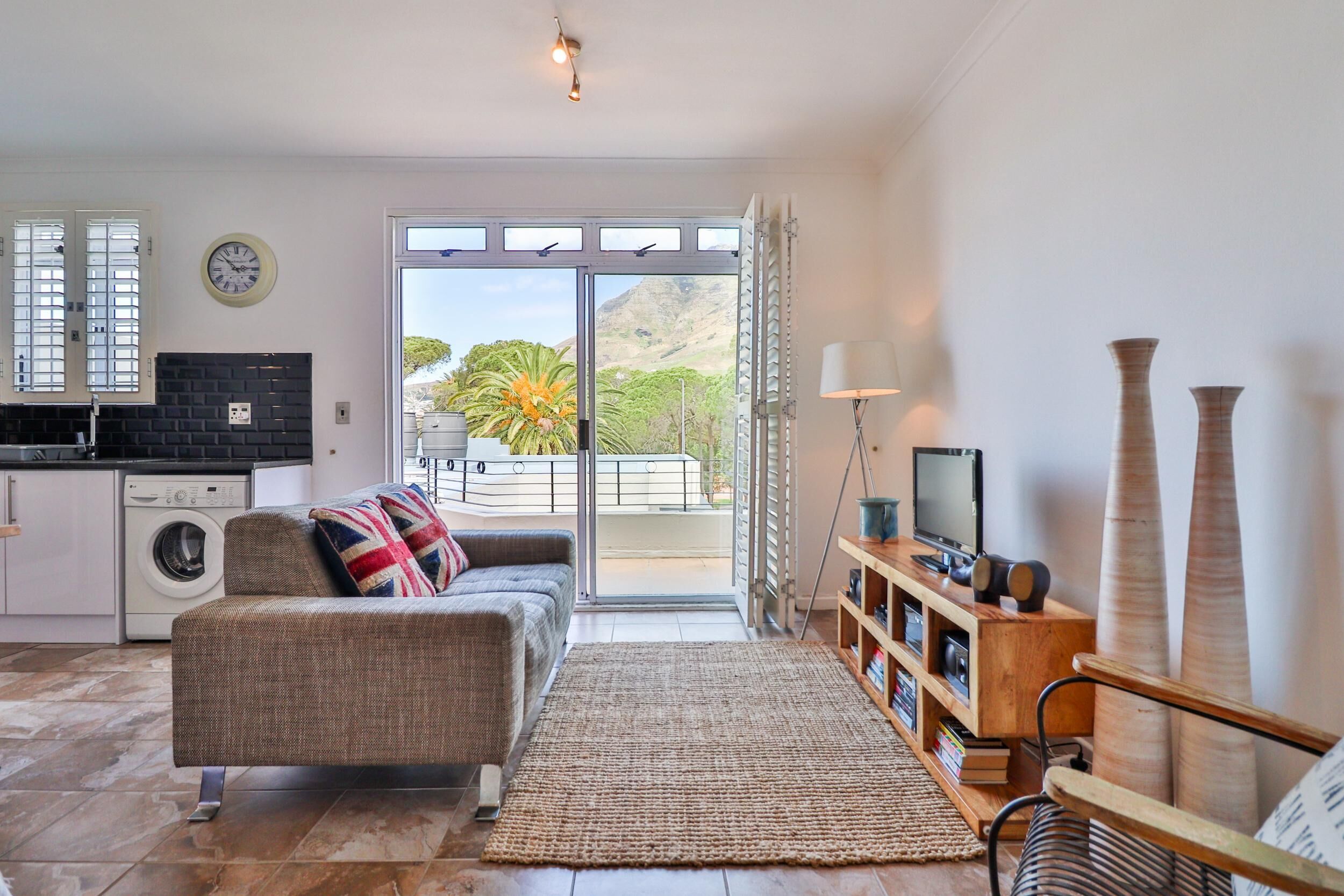 Sala de estar com sofá, mesa de centro, televisão e estante de livros. Há uma porta de vidro deslizante que dá para uma varanda com vista para uma montanha e uma palmeira.