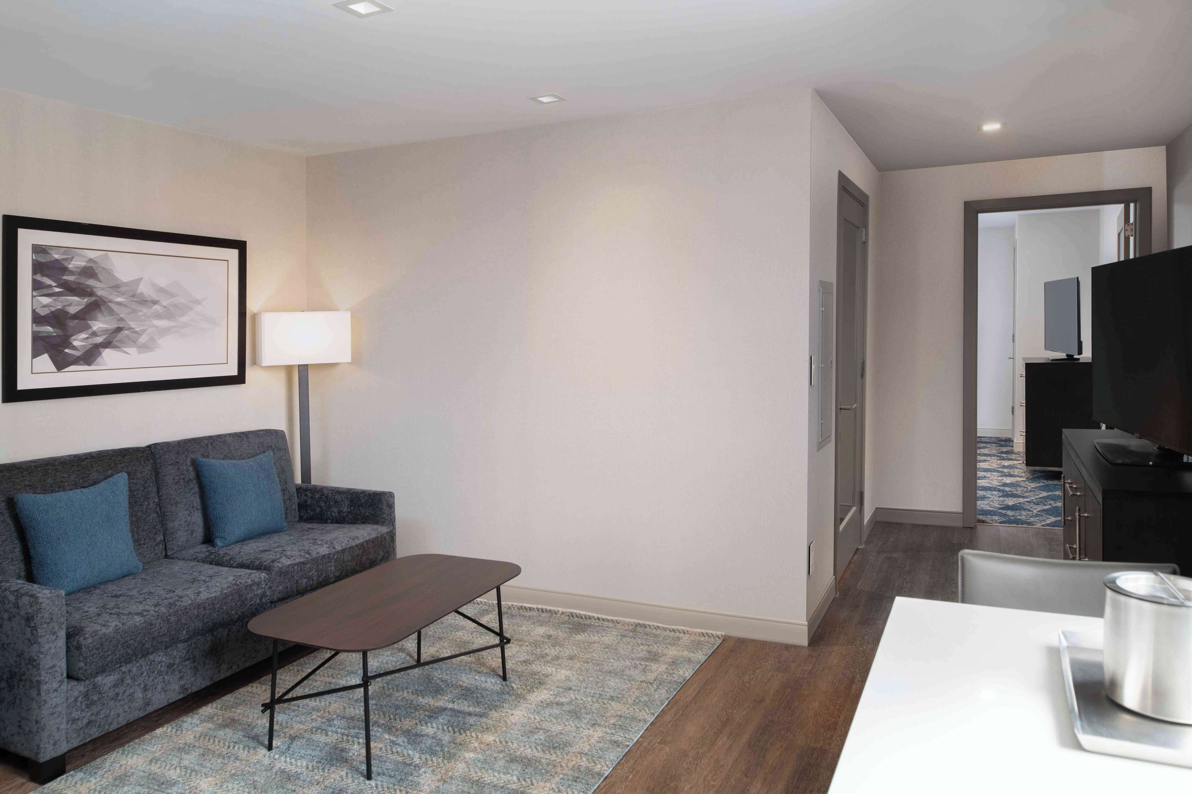 A modern hotel room with a grey sofa, a small table, a lamp, and a kitchen area visible through an open door.