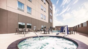 Un jacuzzi rodeado de mobiliario exterior y una piscina, con un edificio al fondo.