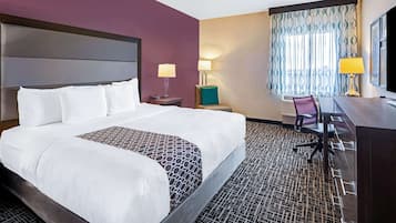 A hotel room with a large bed, a desk with a chair, a nightstand with a lamp, and a window with curtains.