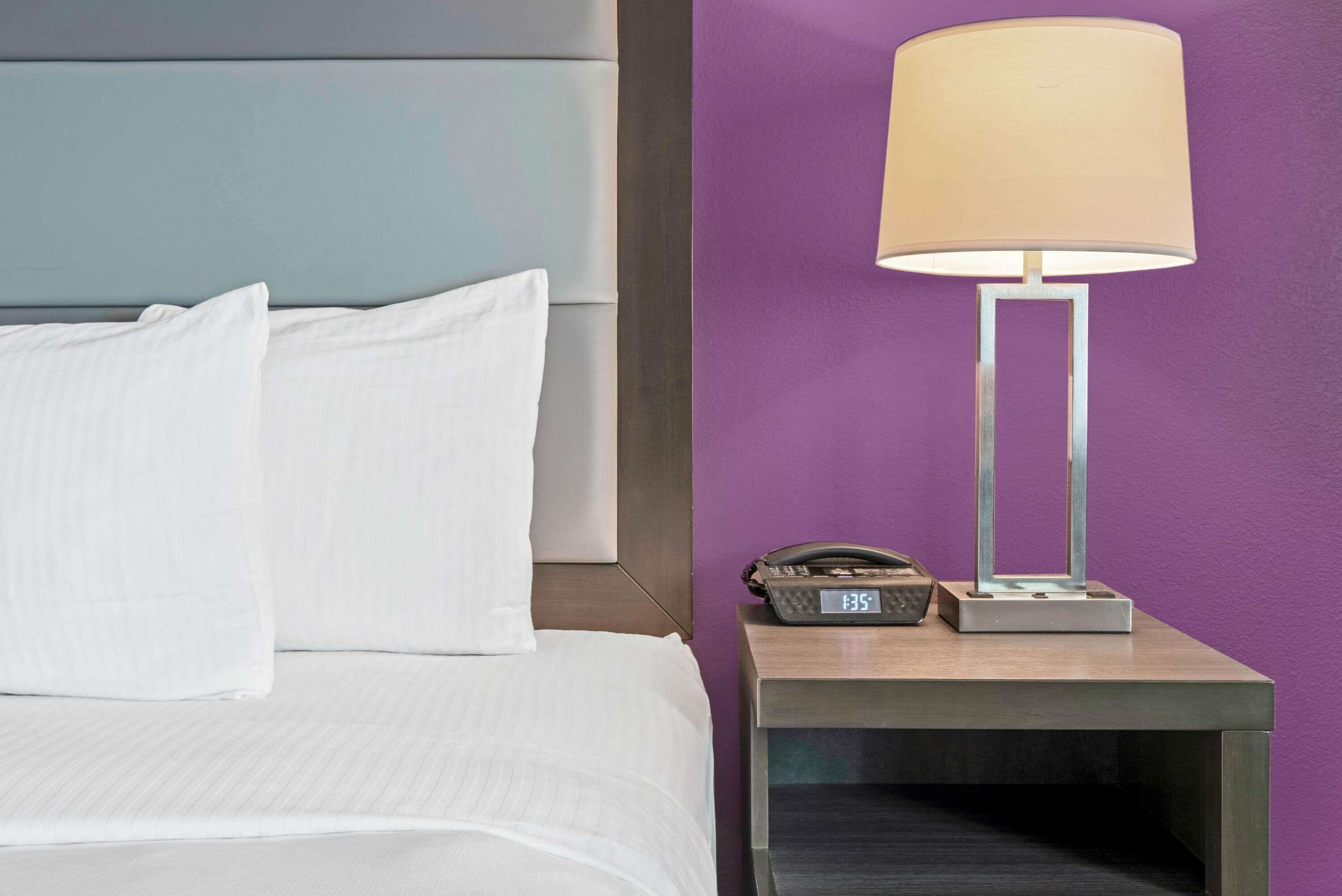 Una cama bien tendida con sábanas y almohadas blancas, una lámpara moderna sobre una mesita de noche y un reloj digital que muestra la hora.