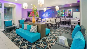 Un moderno vestíbulo de hotel con un sofá azul, sillas color turquesa, una alfombra estampada y una zona de bar con estantes llenos de botellas.
