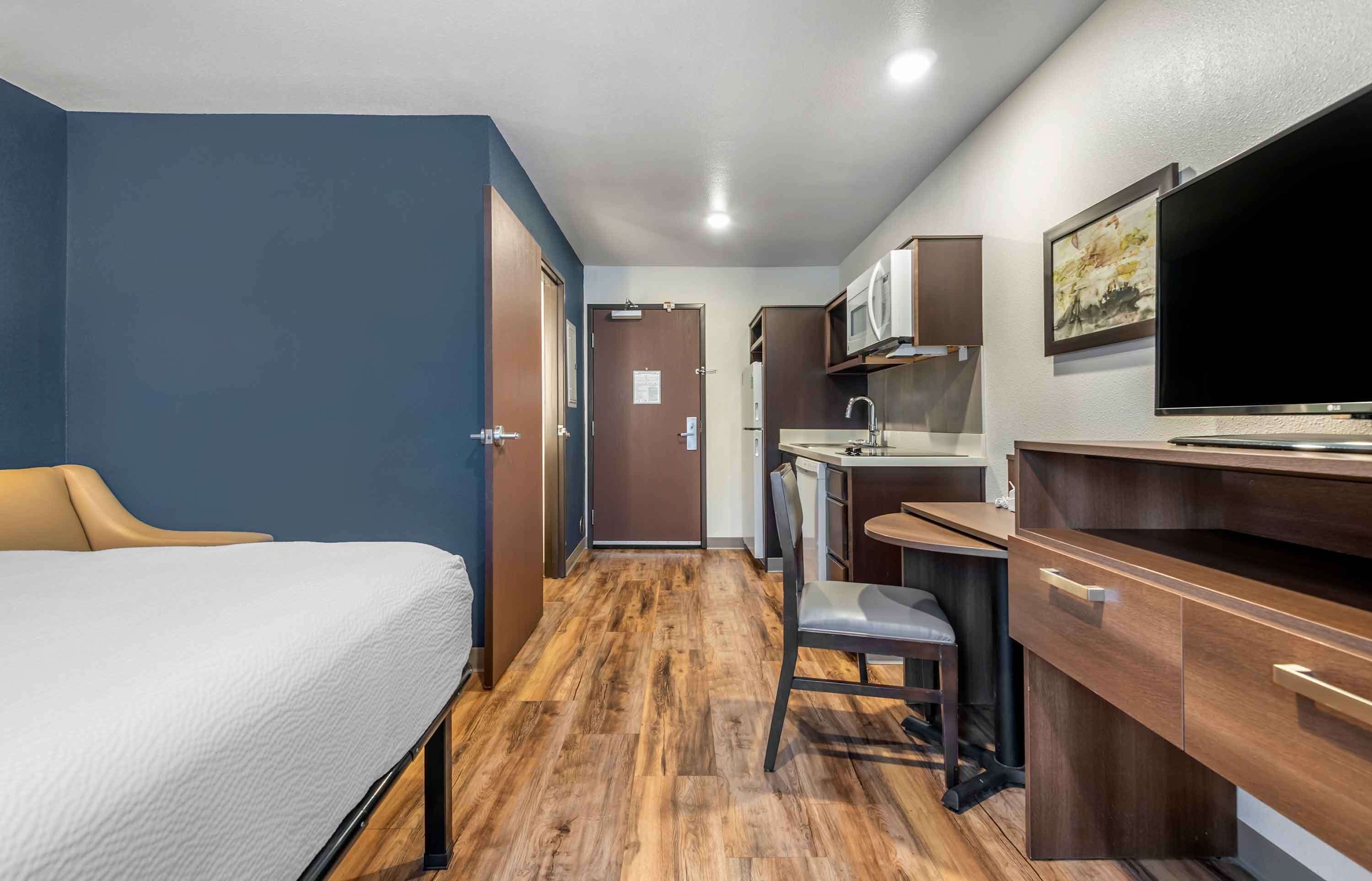 A compact hotel room with a bed, a desk, a chair, a microwave, and a TV.