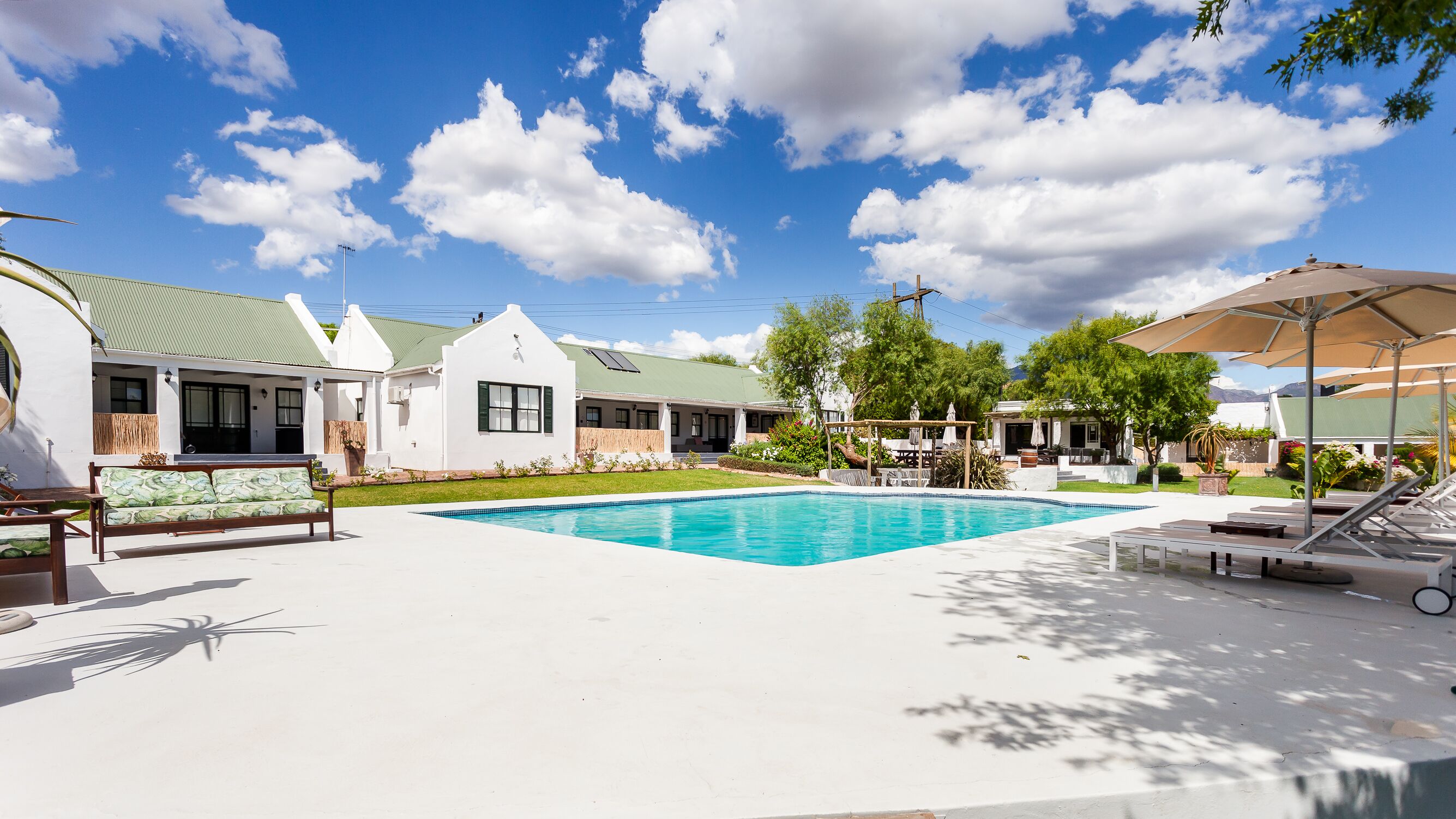 Una piscina con sillones reclinables y sombrillas, rodeada por un edificio blanco con techo verde y un patio cubierto.