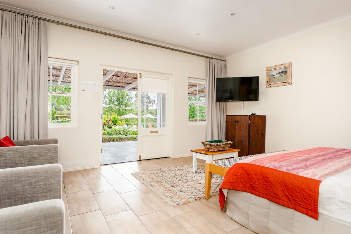 Un dormitorio con cama, televisor, escritorio, silla y vista al jardín por la puerta.