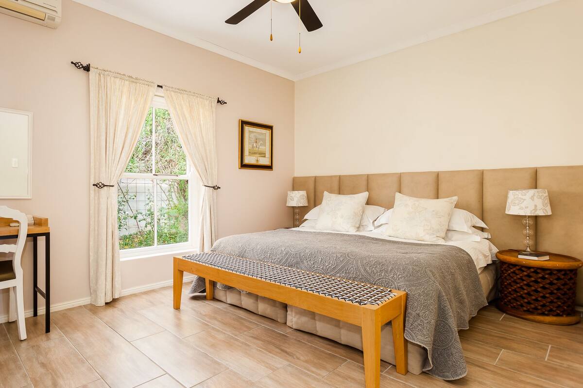 Un dormitorio con cama, lámparas de noche, un banco de madera, un escritorio con silla, una ventana con cortinas y un ventilador de techo.