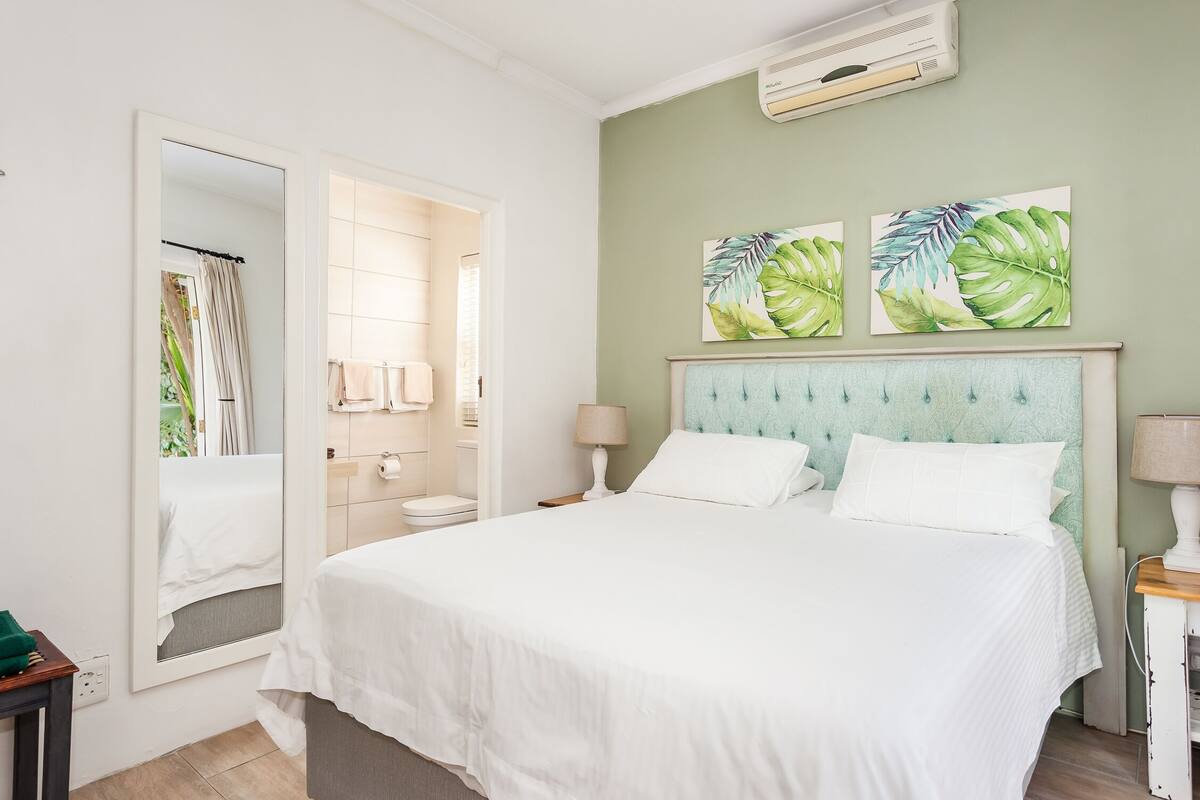 Un dormitorio con cama, mesita de noche, lámpara, espejo y dos cuadros colgados en la pared.