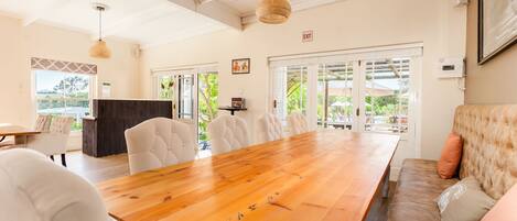 Uma sala de jantar espaçosa com uma mesa de madeira e cadeiras estofadas brancas, grandes janelas com vista para o verde, e um espaço de estar acolhedor com um sofá confortável.