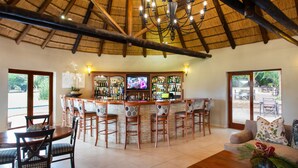 A bar area with a thatched roof, wooden chairs, a television, and a bar stocked with bottles. - Falaza Game Lodge (Hluhluwe)