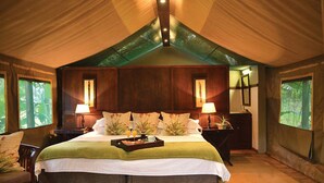 A spacious tent-style room with a large bed, two bedside tables with lamps, and a view of greenery through the windows. - Falaza Game Lodge (Hluhluwe)