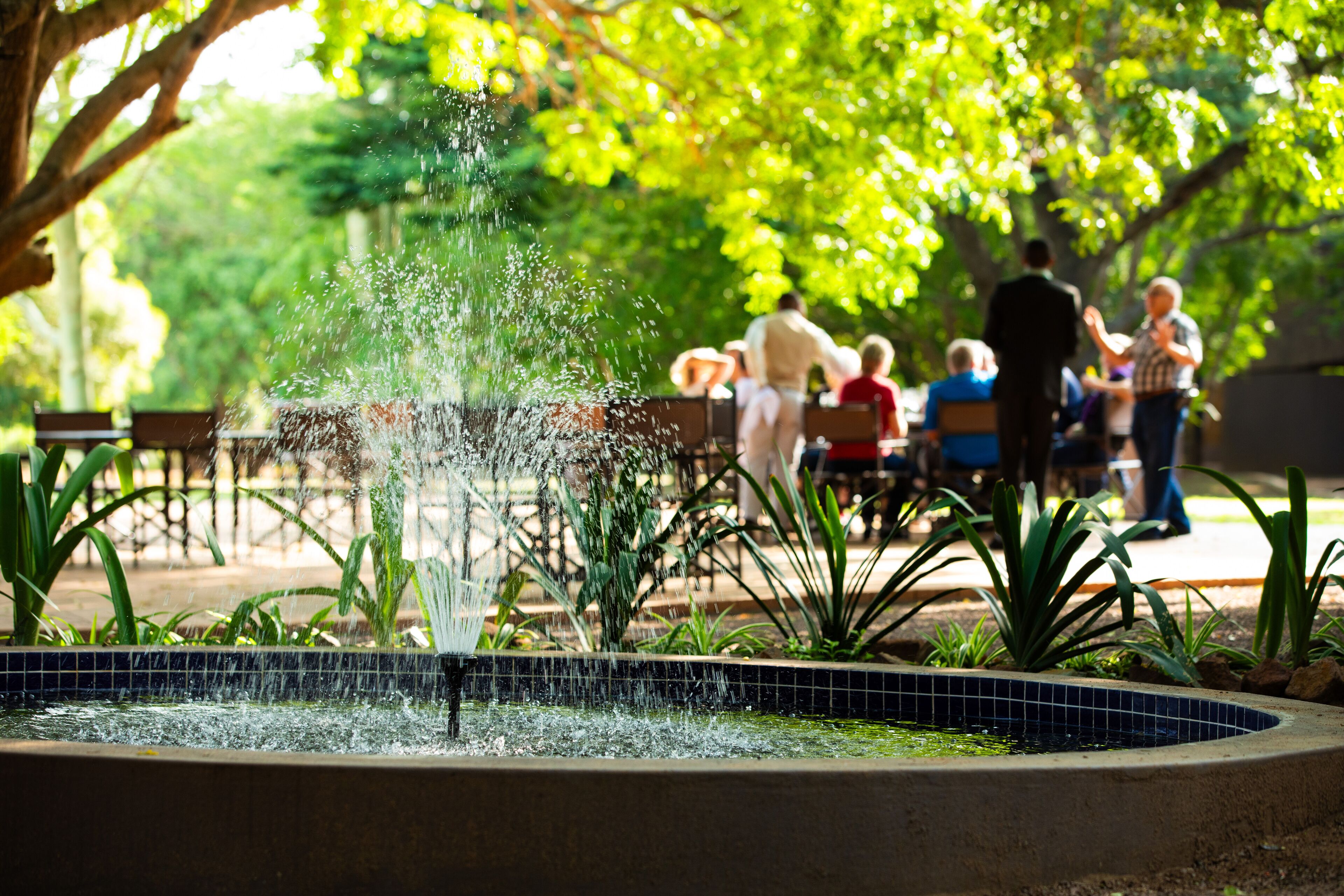 A fountain with water jets in a tiled basin, surrounded by greenery and benches.