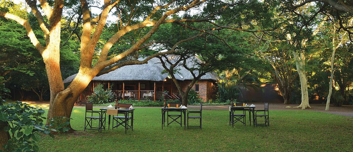 Un espace repas aménagé à l’extérieur avec des tables et des chaises, entouré d’arbres et bordé d’un bâtiment dont le toit est recouvert de chaume.